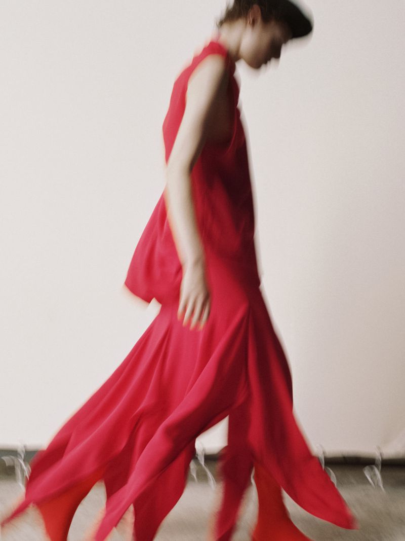 An adult female fashion model wearing a flowing, red dress walks across a light studio backdrop, captured with motion blur.