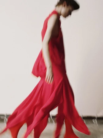 An adult female fashion model wearing a flowing, red dress walks across a light studio backdrop, captured with motion blur.