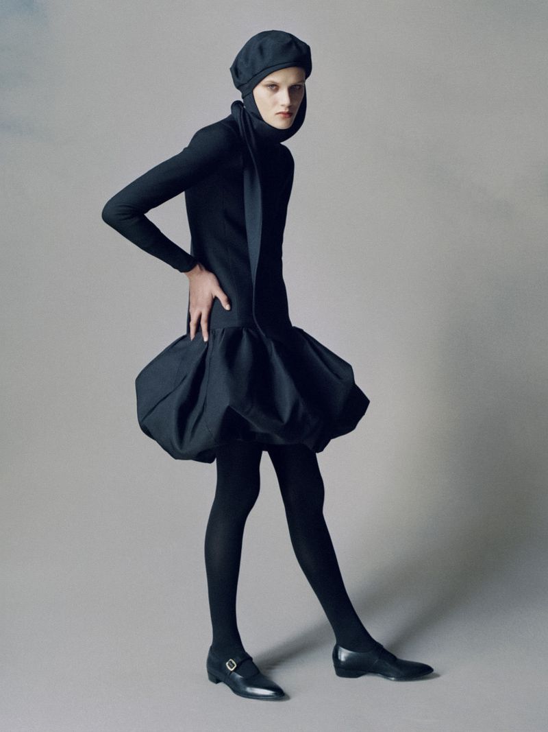 A model poses in a black head covering, top, and bubble dress against a grey background. The outfit is paired with tights and flats.