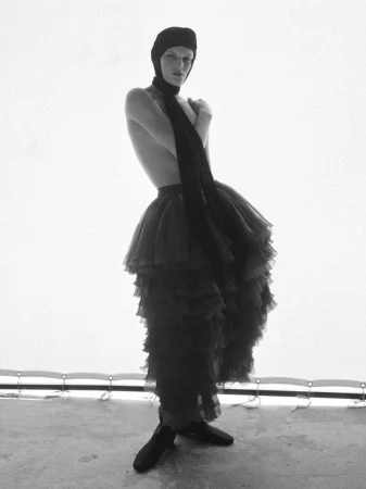 Monochrome portrait of a gender-nonconforming person in a tulle skirt, headdress, and ballet shoes posing against a white backdrop.