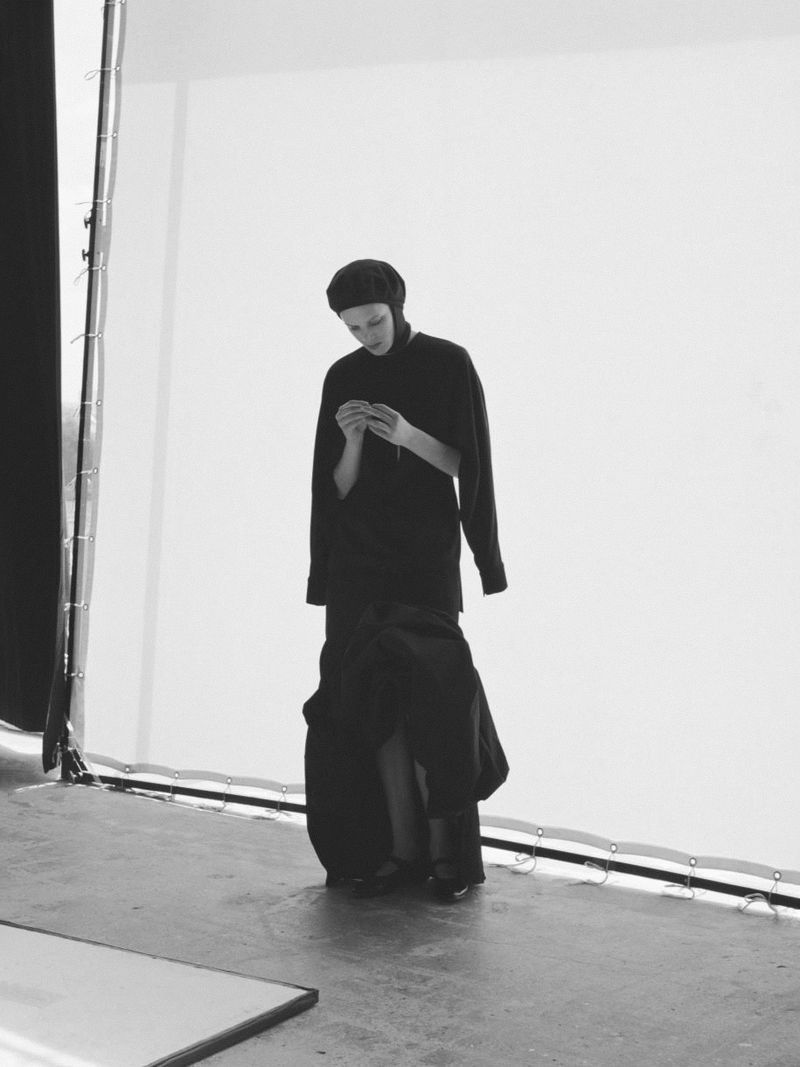 Black and white, medium shot of a young adult female in a dark outfit and beanie, standing in a photo studio, looking down at her hands.
