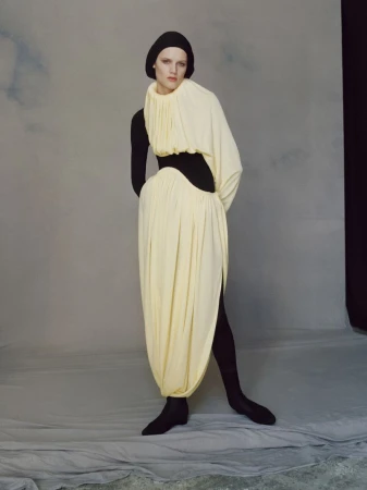 Model Noor Khan wearing a sculptural pleated pale yellow draped dress over a black hood and bodysuit against a grey studio backdrop.