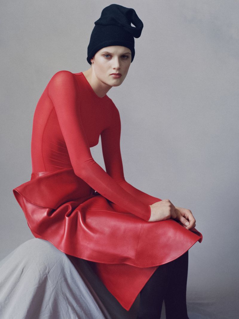 A woman with fair skin is seated, wearing a long-sleeved red dress with a leather skirt and a black beanie against a grey background.