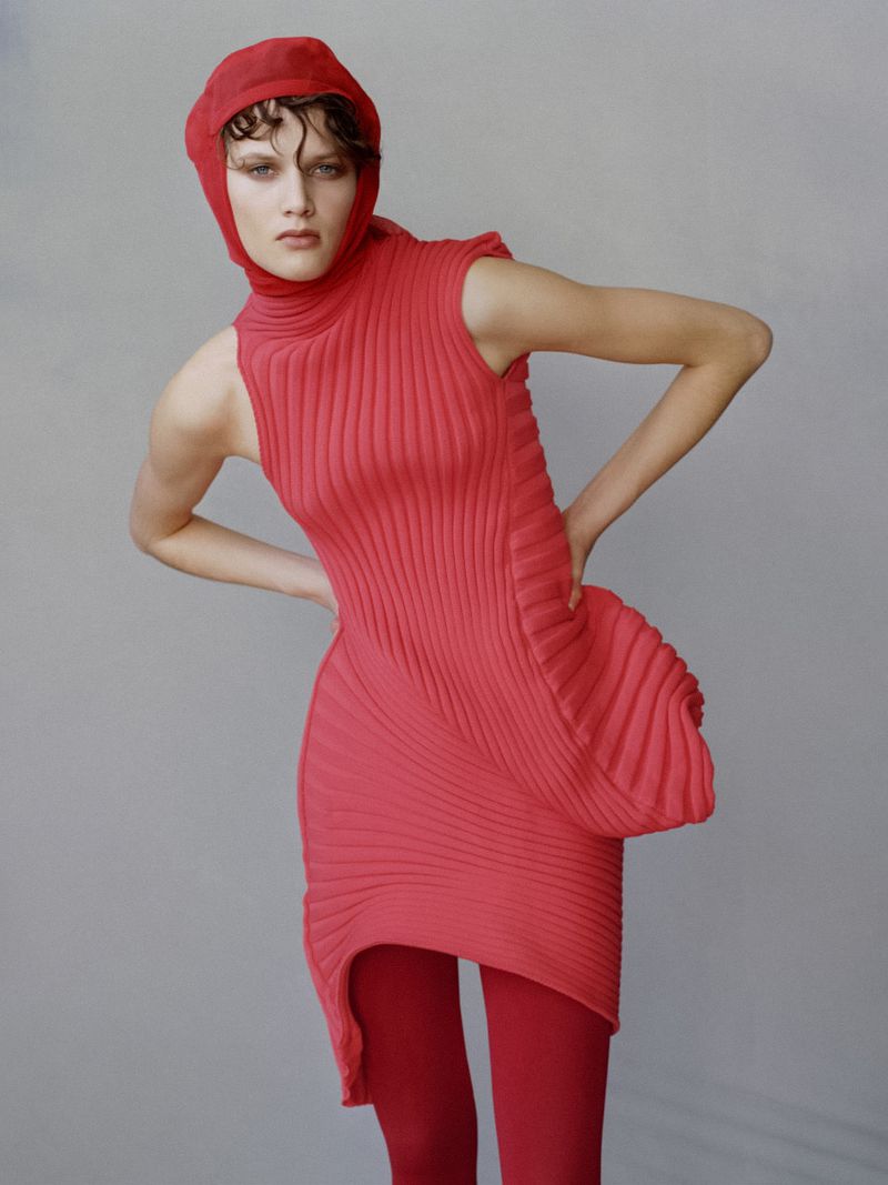 Fashion model poses in a studio wearing a red knit dress, head covering, and tights against a neutral gray background in a studio setting.