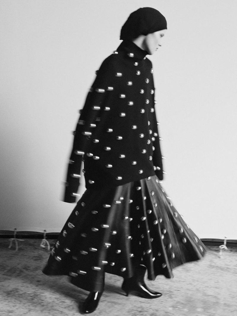 Monochromatic studio shot of a woman in a black studded sweater and full skirt, paired with heeled boots and a matching head covering.