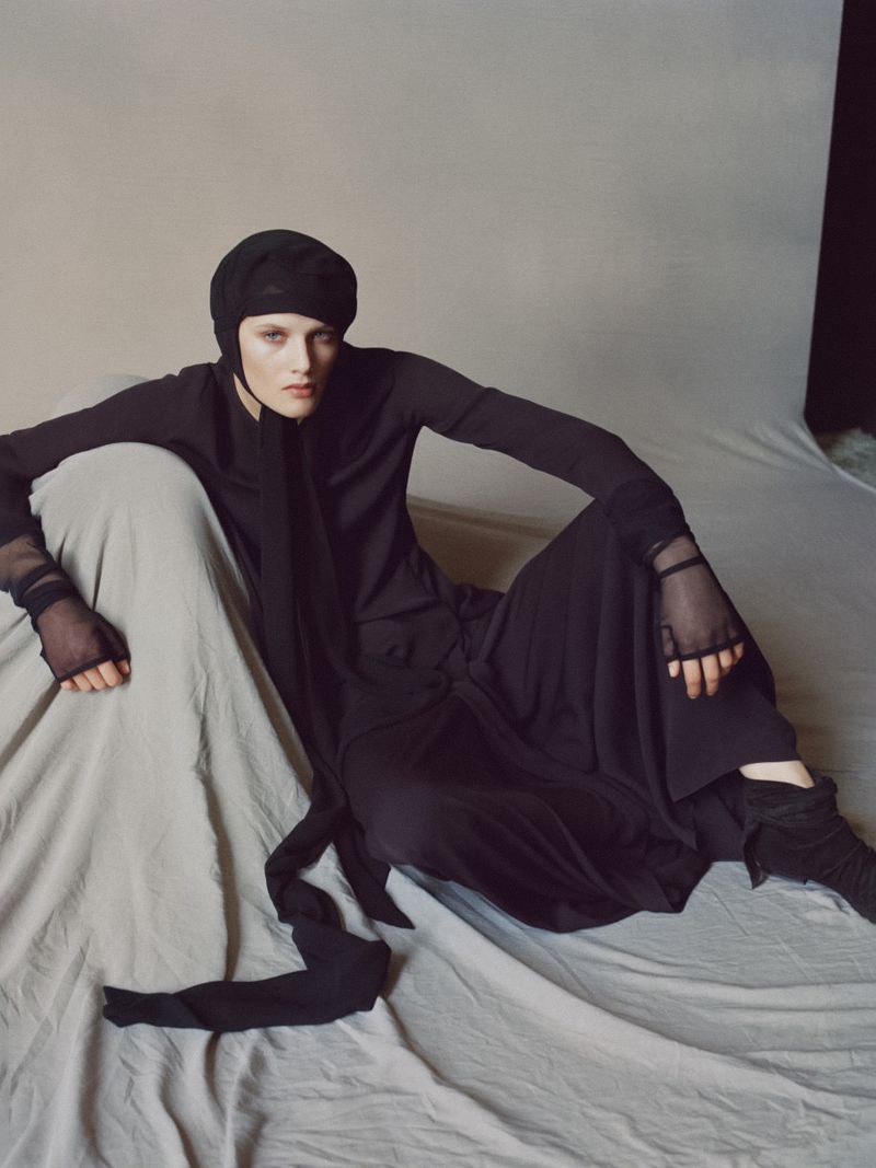 A model in a black outfit sits on a rumpled grey fabric, styled with a head covering and mesh gloves in a studio fashion shoot.