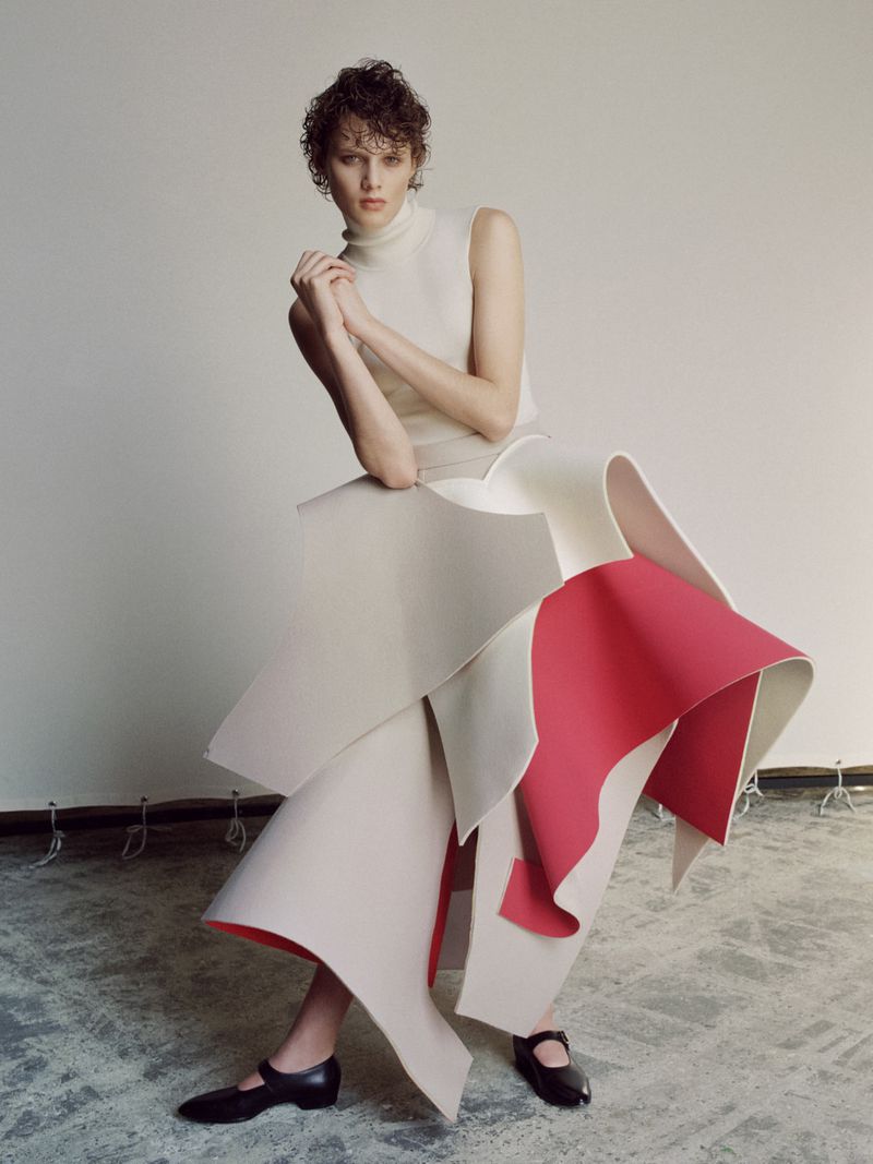 Fashion editorial image of a young adult woman in a high-neck top and sculptural layered skirt posing against a plain studio backdrop.