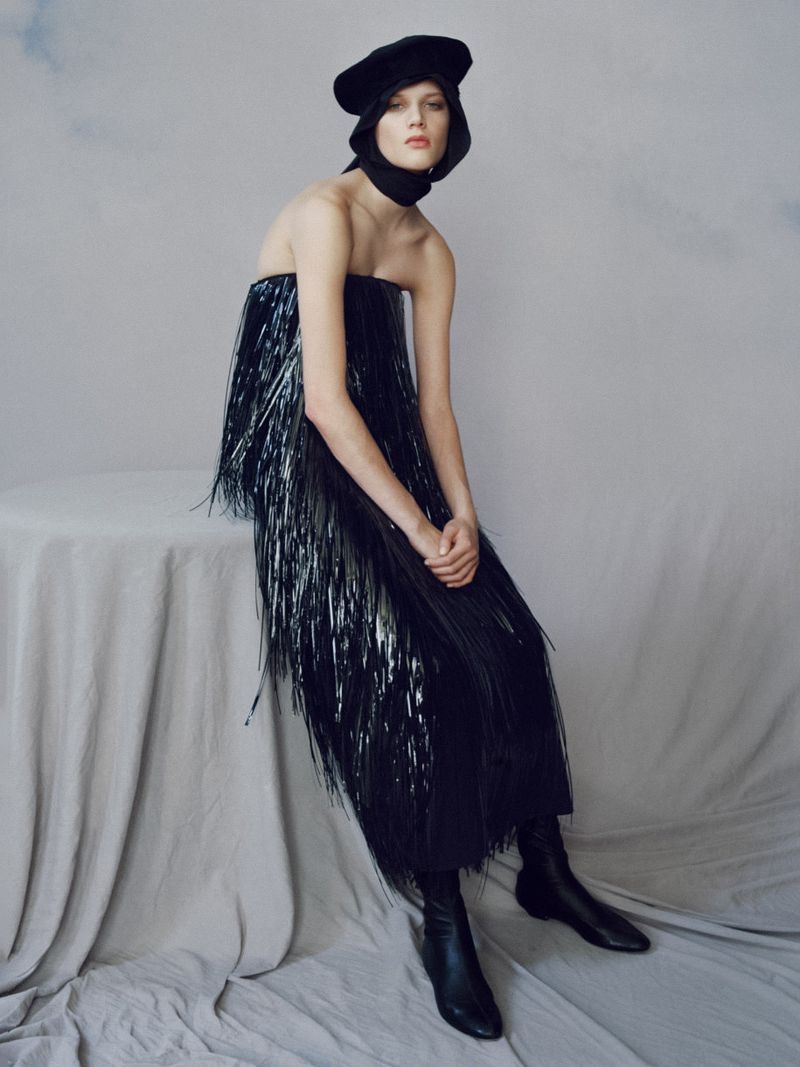 Fashion shot of a woman in a dark, fringed dress, seated on a draped table, wearing a black beret and boots, against a cloud-painted backdrop.