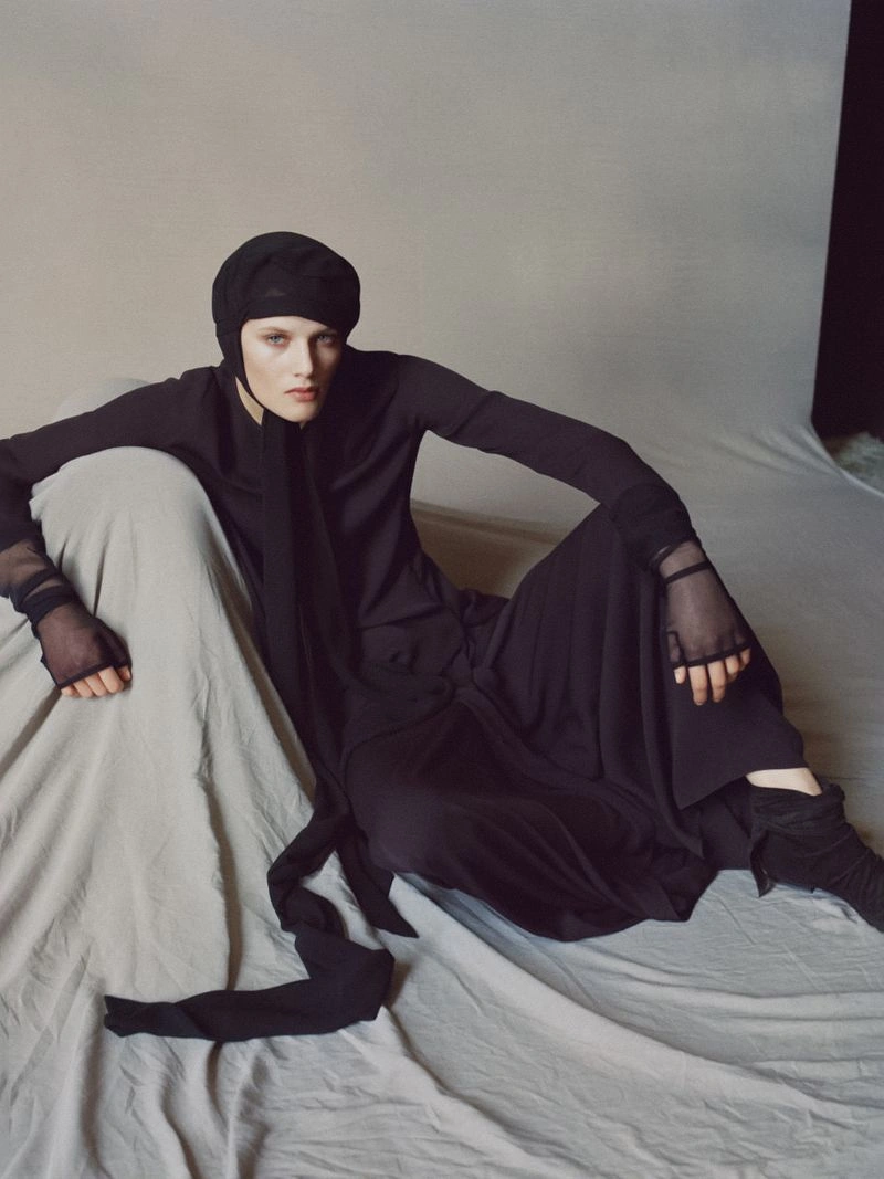 Model Noor Khan wearing a black hooded headscarf, long-sleeve garment, and sheer gloves, seated on draped fabric in a studio setting.