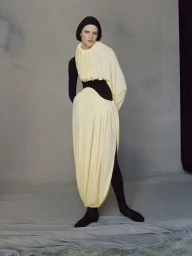 Model Noor Khan wearing a sculptural pleated pale yellow draped dress over a black hood and bodysuit against a grey studio backdrop.