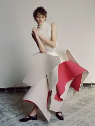 Model Noor Khan poses in a cream sleeveless turtleneck and an architectural layered pink and beige skirt in a studio setting.