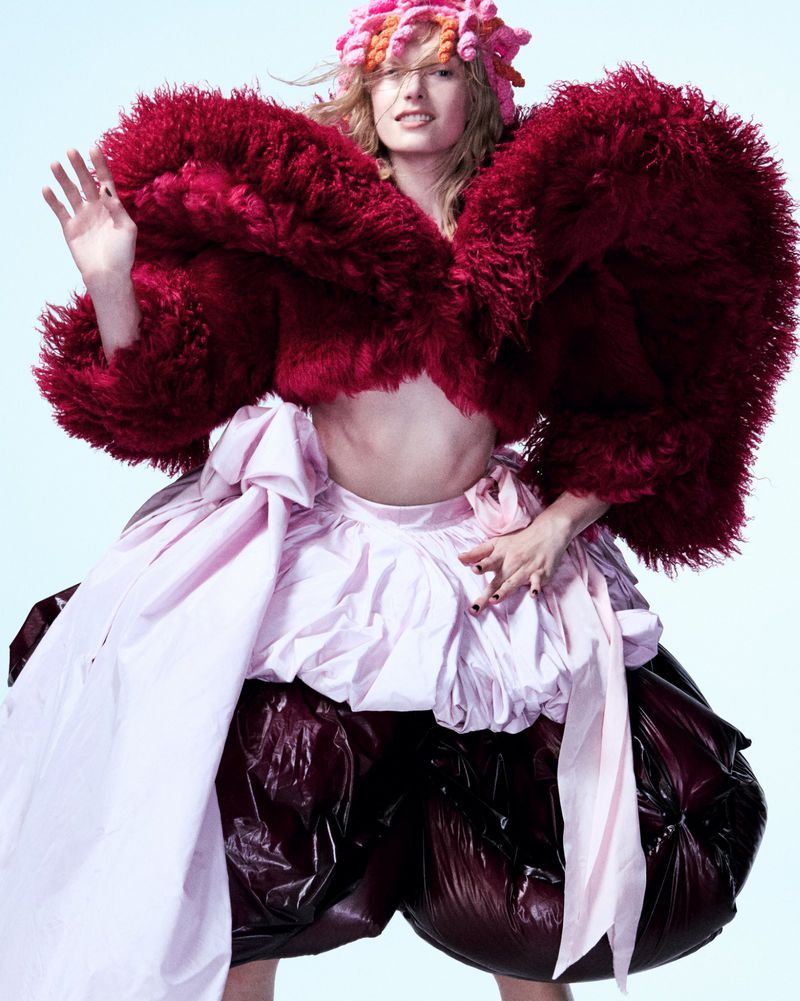 Editorial fashion shot of a posing woman wearing a flamboyant red faux fur bolero jacket, pink draped skirt, and a pink knitted hat.
