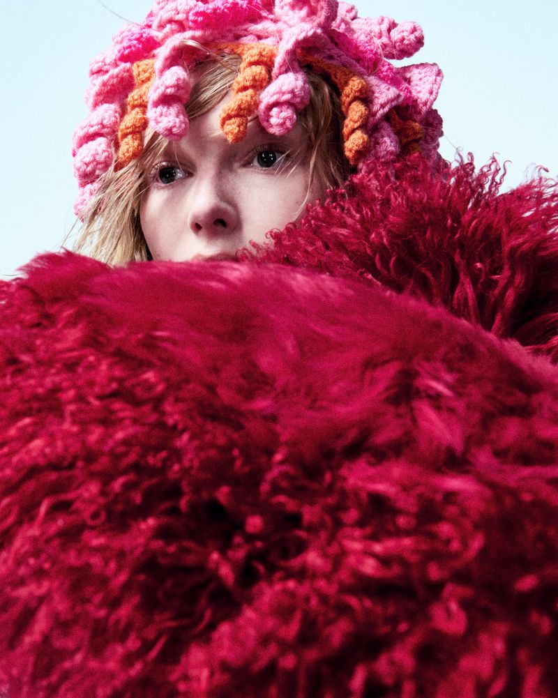 Close-up studio shot of a woman wearing a vibrant pink and orange crocheted hat and a bright red faux fur collar, gazing directly at the viewer.