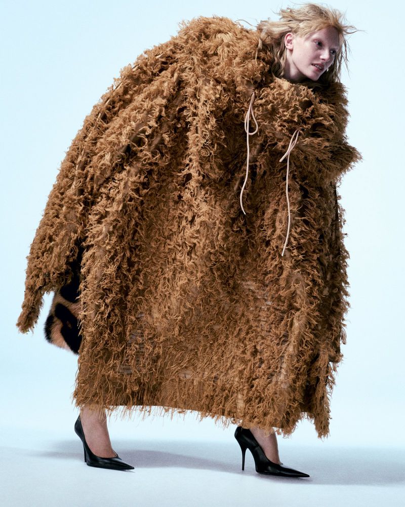 Woman in a large brown textured fashion coat with black high heels posing in a light studio environment. Fashion editorial shot.