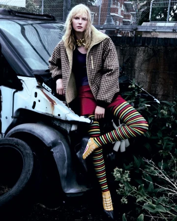 A blonde model poses in front of a wrecked car, wearing a plaid jacket, red skirt, striped leggings, and yellow crocodile shoes in an outdoor urban setting.