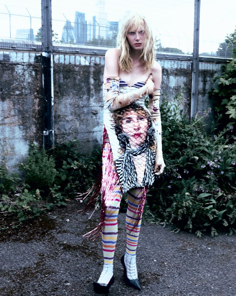 Young adult woman poses in patterned dress with pink fringe outdoors, urban background, showcasing fashion and visual artistry.