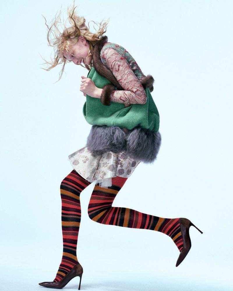 Fashion shot of a young adult woman leaping on a light blue backdrop, wearing a green jacket, mini skirt, patterned tights, and heels.
