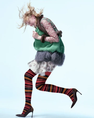 Fashion shot of a young adult woman leaping on a light blue backdrop, wearing a green jacket, mini skirt, patterned tights, and heels.