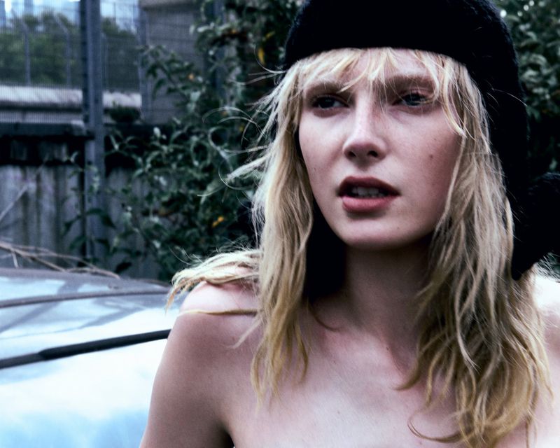 Portrait of a fair-skinned woman with blonde hair and a dark cap, captured outdoors against a blurred background of trees and structures.