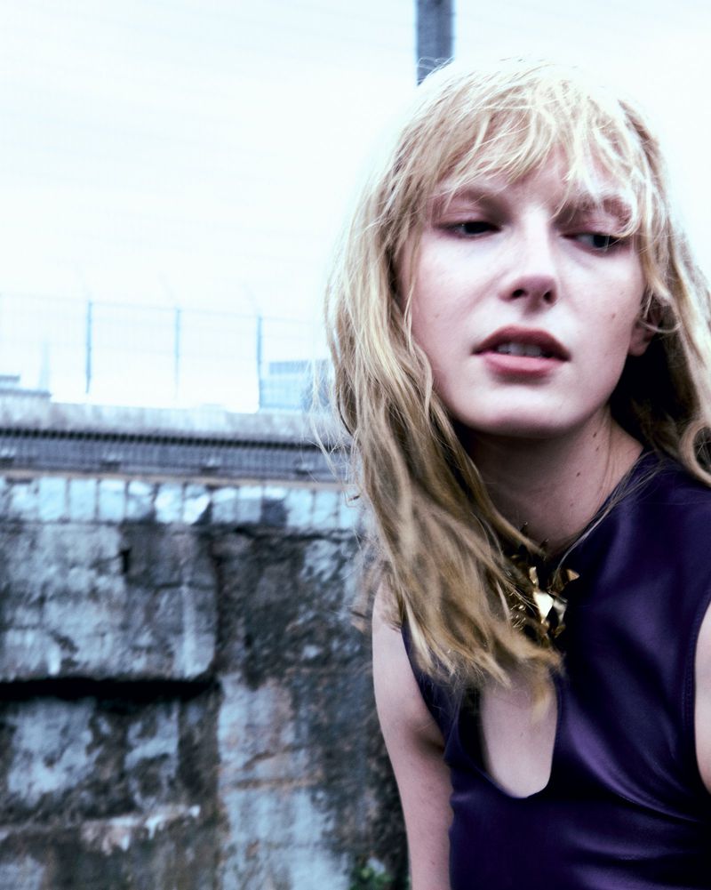 Blonde woman in a purple top and gold necklace poses outdoors against a stone wall and blurred buildings in an overcast daytime setting.