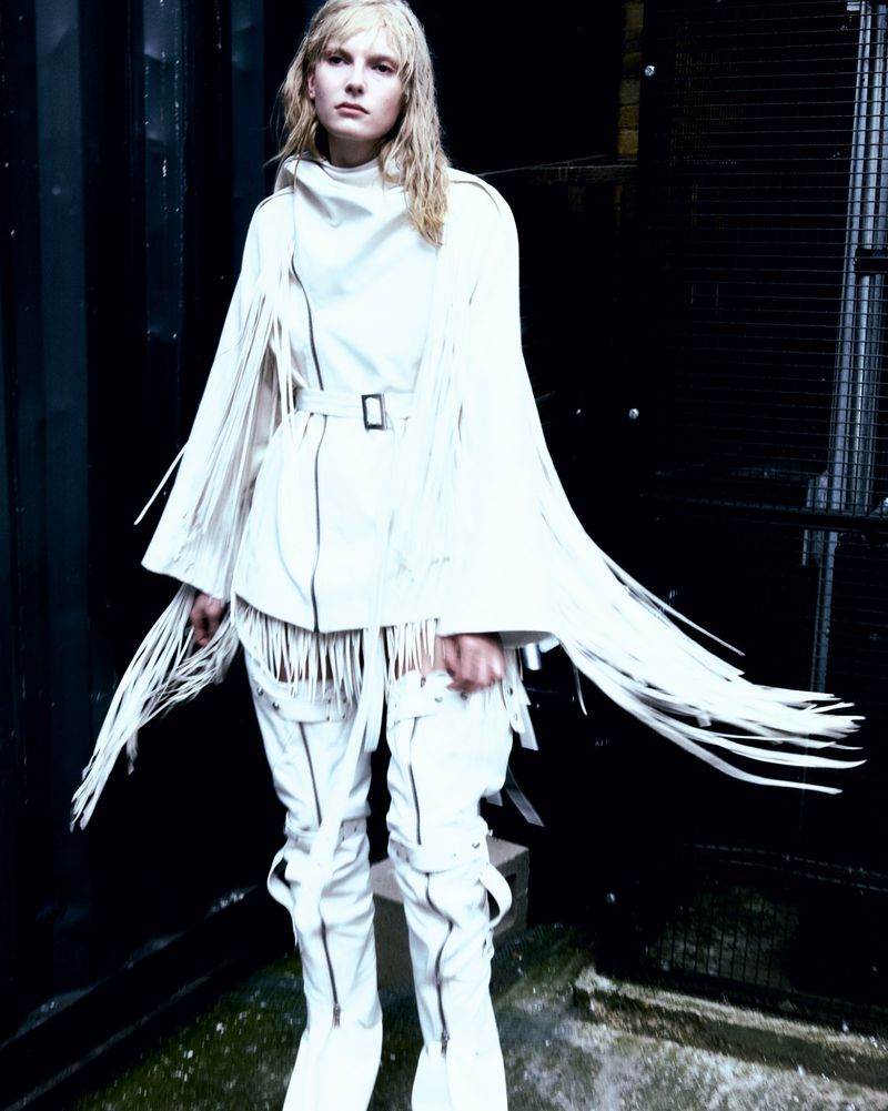 A woman in a white fringe outfit stands against a dark wall, showcasing edgy fashion with a stark contrast and unique textures.