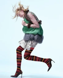 Model Alex Consani mid-stride wearing a green sleeveless top, patterned skirt, and striped tights against white