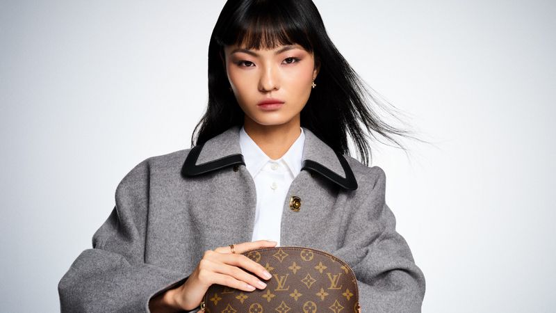 Asian woman in gray coat holds brown Louis Vuitton handbag in a studio setting with white background, showcasing fashion and beauty.