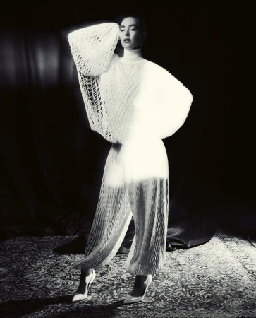A woman in a white textured outfit poses on a patterned carpet against a dark background in a fashion-style, studio shot.