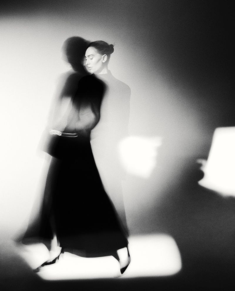 Black and white portrait of a woman in a blurred walking pose. The image features dynamic motion blur and bright spot lighting.