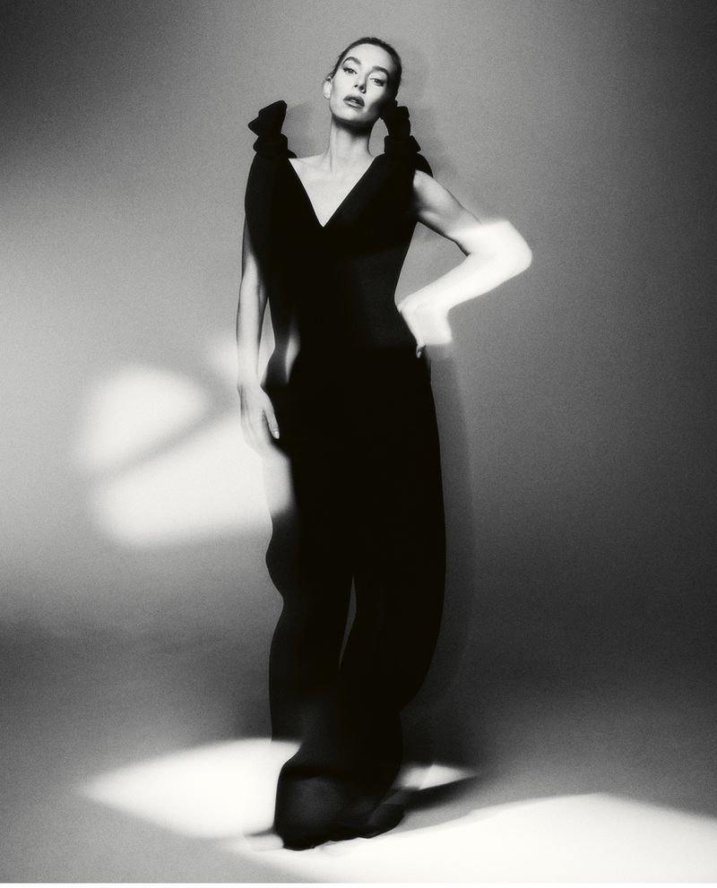 A woman poses in a studio wearing a black dress with bow details on the shoulders, highlighted by monochrome lighting.