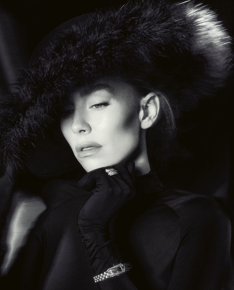 Monochrome portrait of a woman with a feathered hat, gloved hand to face, and a vintage wristwatch in a dark, soft-lit indoor setting.