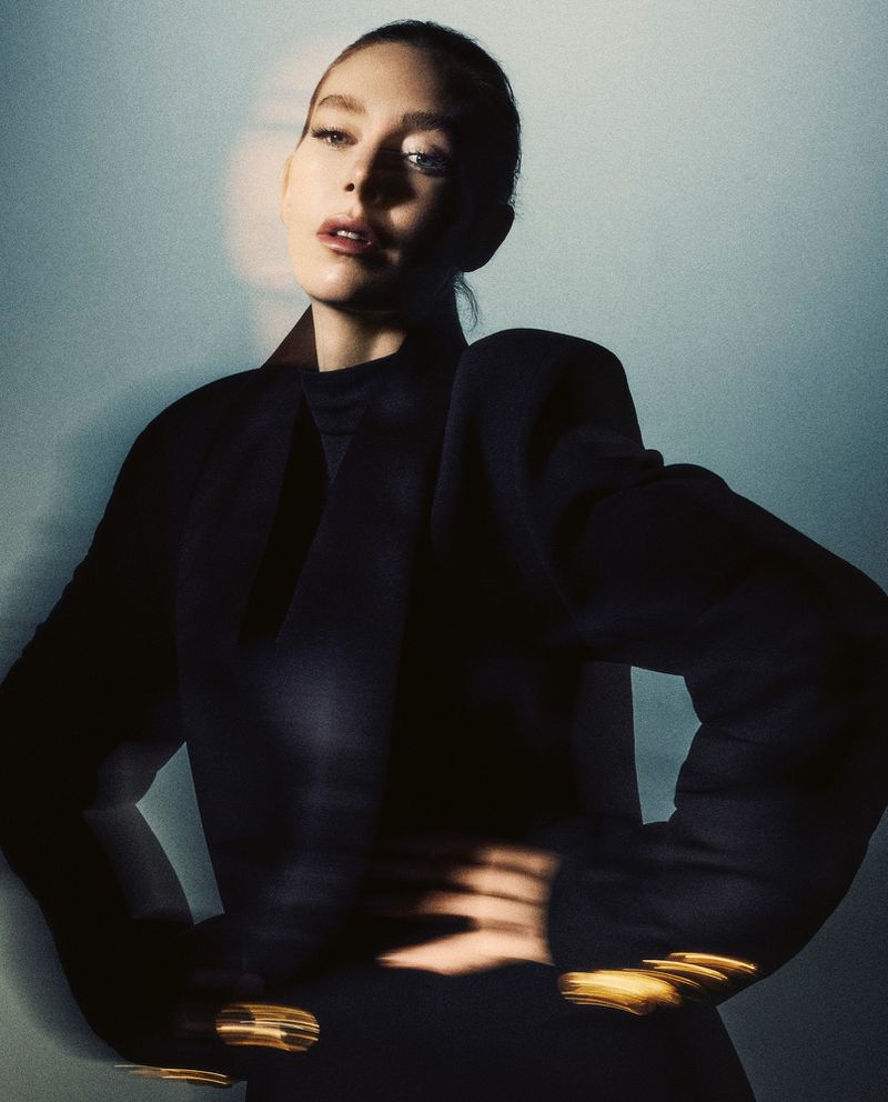 Indoor portrait shot of a stylish woman in a dark designer blazer posing with hands on hips against a blue-grey gradient background.