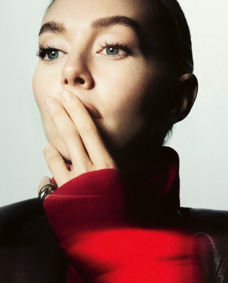 Close-up studio portrait of a young adult woman with hand near face, wearing a red turtleneck sweater and leather jacket, on a white background.