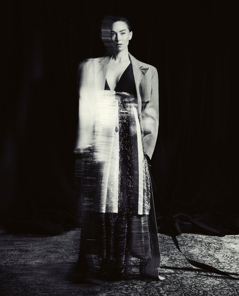 Monochrome full shot of a woman in a gray blazer, black top, and long skirt with a blurred effect, standing indoors against a dark backdrop.
