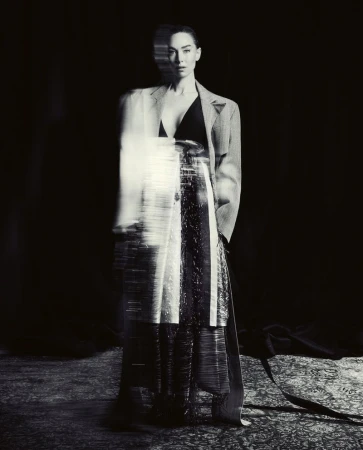 Monochrome full shot of a woman in a gray blazer, black top, and long skirt with a blurred effect, standing indoors against a dark backdrop.
