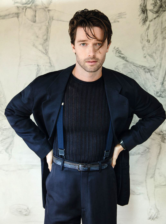 Portrait of a handsome adult man wearing a black sweater, blue suspenders, and a dark blue jacket posing indoors against an abstract background.