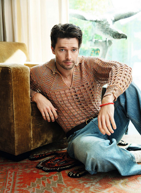 A man with dark hair sits on a carpet near a tan sofa, wearing jeans and a light brown open-knit shirt, creating a relaxed and casual atmosphere.