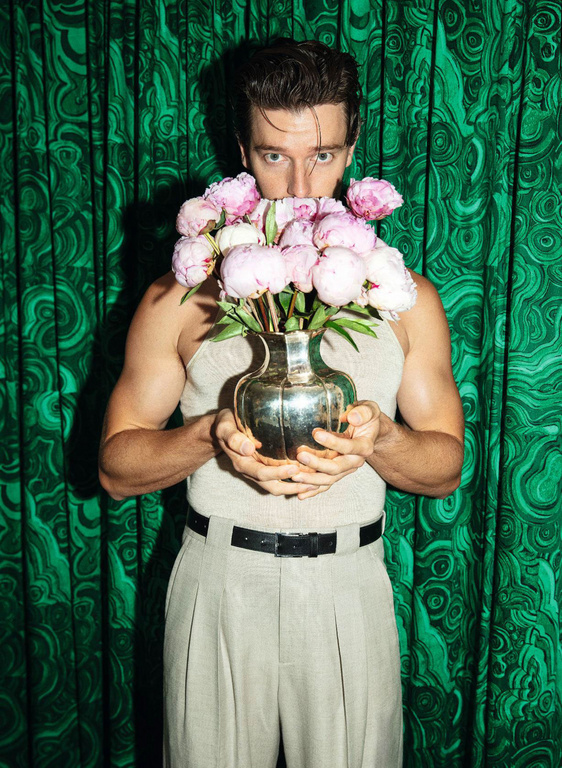 Man in tank top holds a vase of pink peonies against a green patterned background. Male portrait with flowers and a studio setting.
