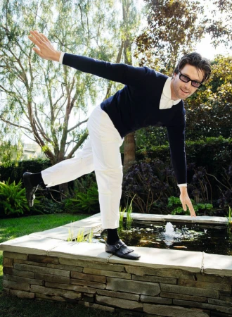 Man strikes balancing pose on stone ledge near a pond in an outdoor garden setting with grass and trees, wearing casual attire.