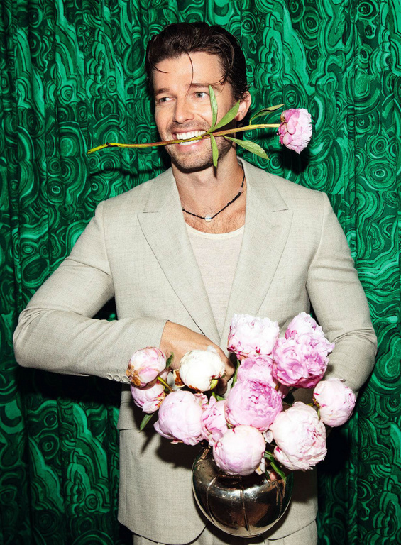 Smiling man in a light-colored suit holds a vase of pink peonies, with a green malachite patterned background, presenting a refined aesthetic.