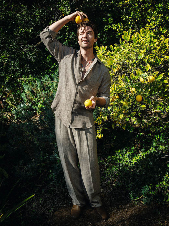 Adult male in casual linen clothing holding lemons, smiling, in a sunlit outdoor setting with a lemon tree and lush green foliage.
