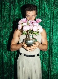 Patrick Schwarzenegger holding a vase of pink peonies in front of a green patterned curtain for Esquire Germany