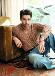 Patrick Schwarzenegger seated on the floor leaning against an armchair wearing a patterned knit top and blue jeans