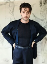 Patrick Schwarzenegger wearing a black blazer and trousers with blue suspenders against a sketched wall backdrop