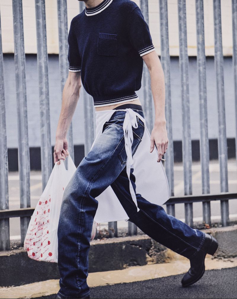 Adult male fashion model in a navy blue knit top, light-wash jeans, and black boots walks in an urban environment carrying a plastic shopping bag.
