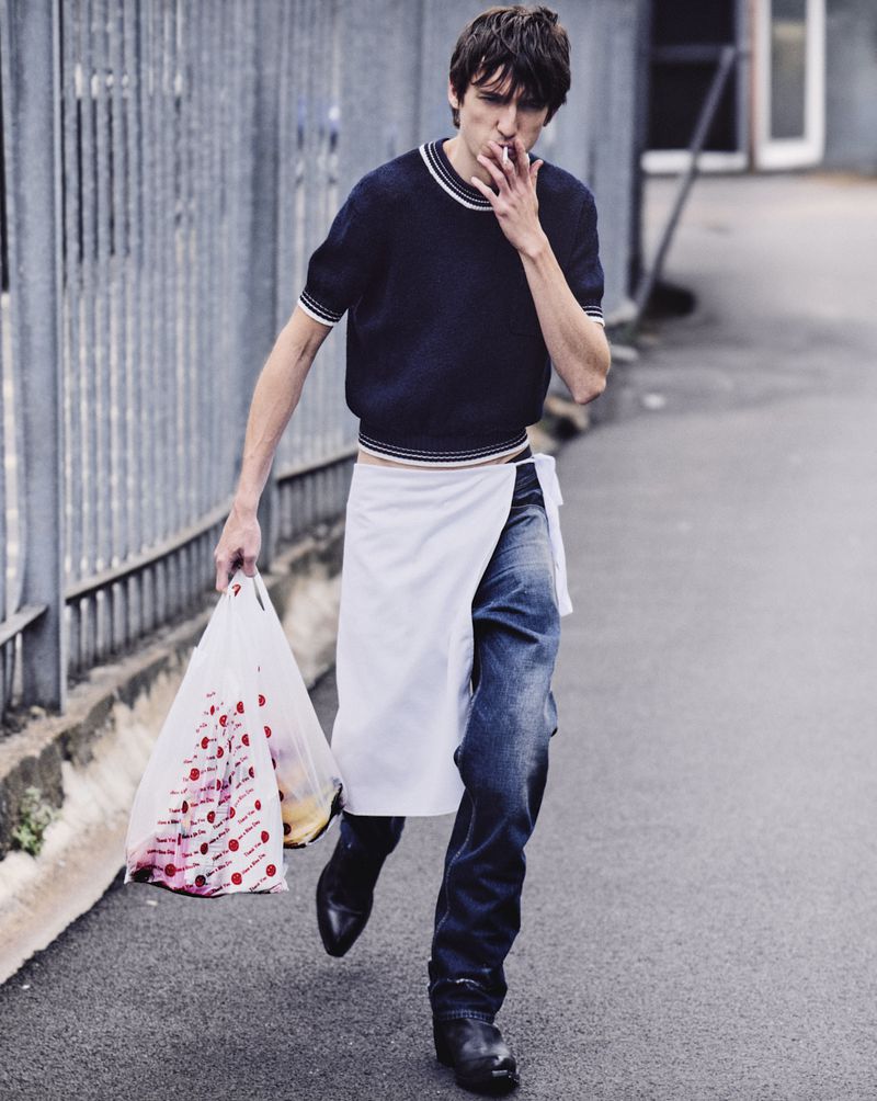 A slender man walks on a street, smoking and carrying a plastic bag with red polka dots, wearing a white apron and blue jeans.