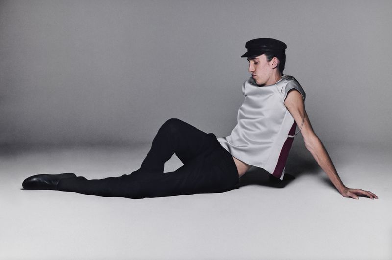 A young man wearing a black cap, silver sleeveless top, and black trousers reclining on a grey studio floor.
