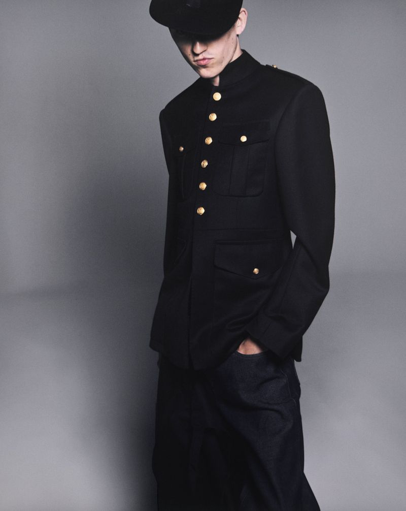Young adult in fashion portrait wearing a black jacket with gold buttons and a black cap against a grey studio backdrop.