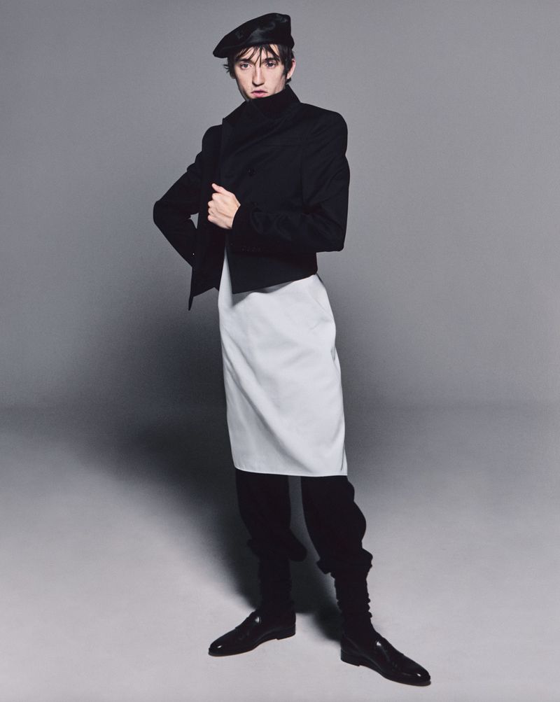 A fashionable adult stands in a photo studio wearing a beret, black jacket, white apron, and black trousers and shoes, against a gray background.