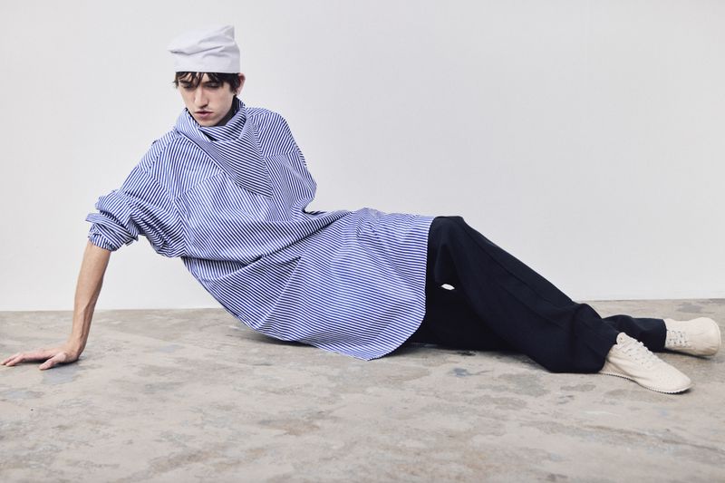 Adult male fashion model poses lying down wearing a striped shirt, dark pants, white shoes, and white cap against a white background.
